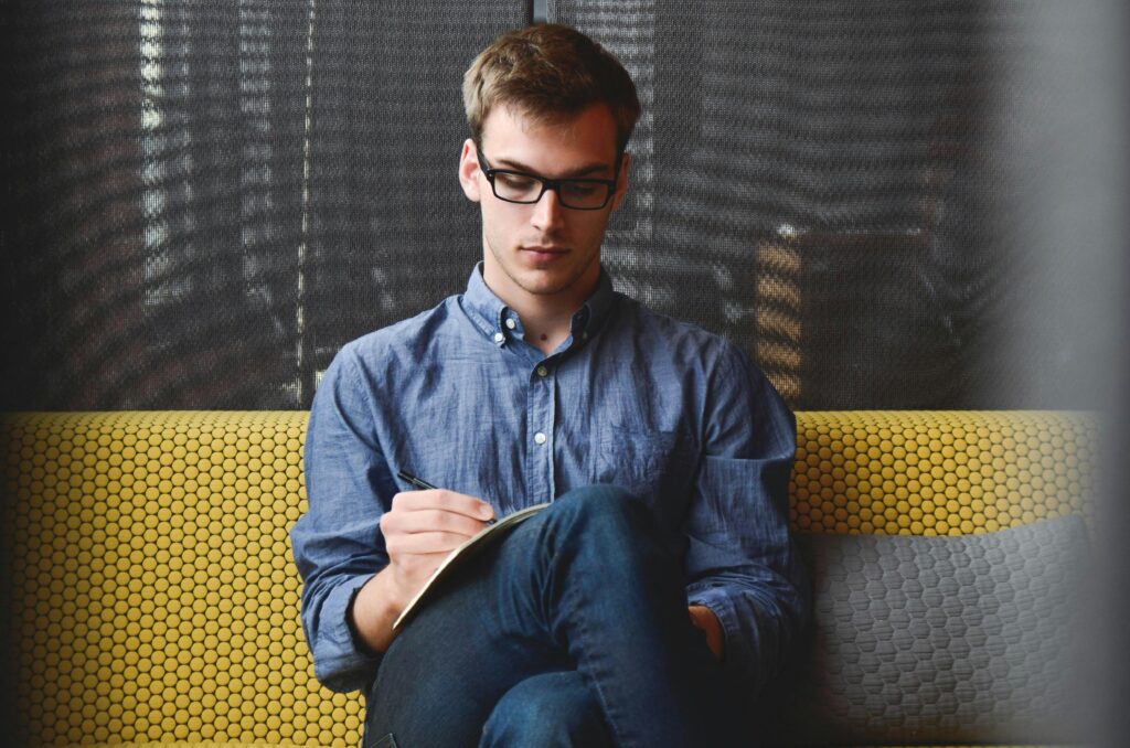 entrepreneur startup start up man 39866 39866 A young man in glasses writes in a notebook while sitting on a stylish couch indoors.